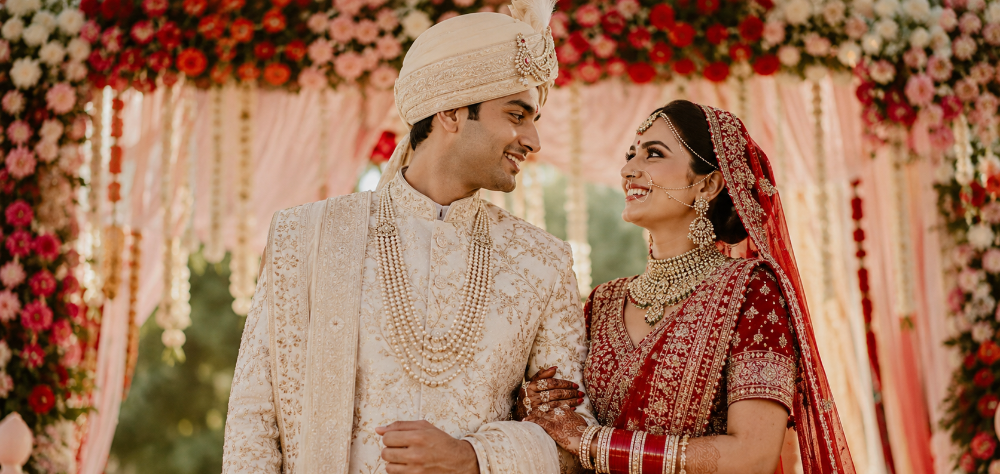Indian groom in an ivory rented sherwani and pearl necklace smiling at his bride