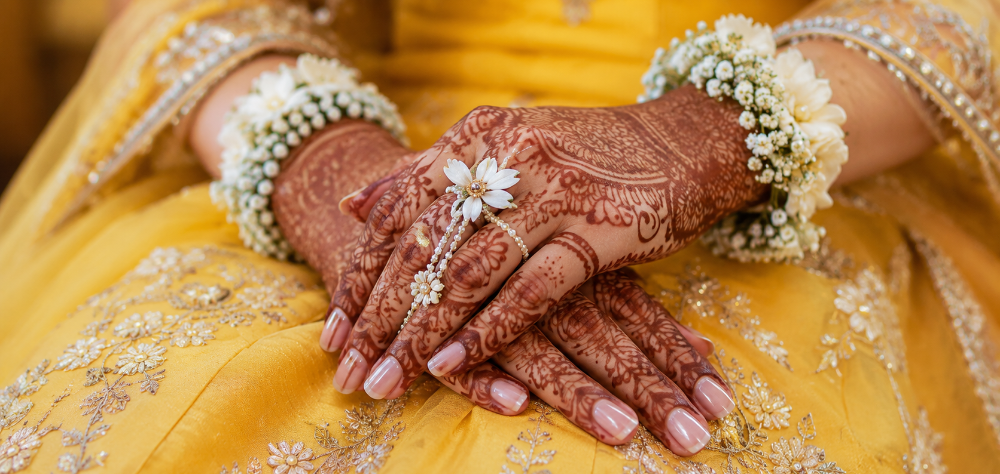Close up of a bride's henna hands and floral jewelry resting on a yellow silk lehenga