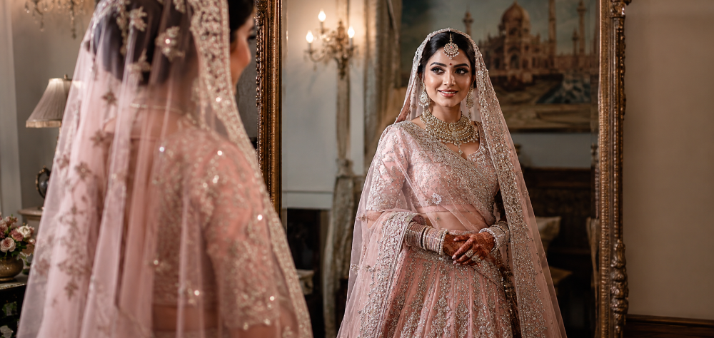 Modern Indian bride admiring her pastel-pink designer lehenga in a vintage mirror