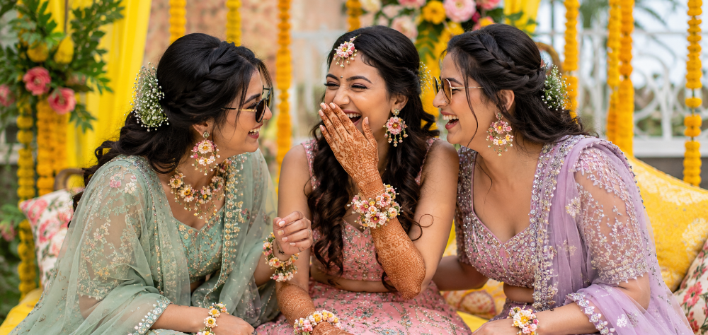 Three bridesmaids wearing coordinated pastel designer lehengas laughing at a Mehndi ceremony